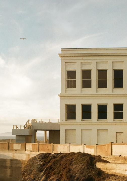 A white building with multiple windows stands on a cliff overlooking the ocean, with a bird flying in the sky.