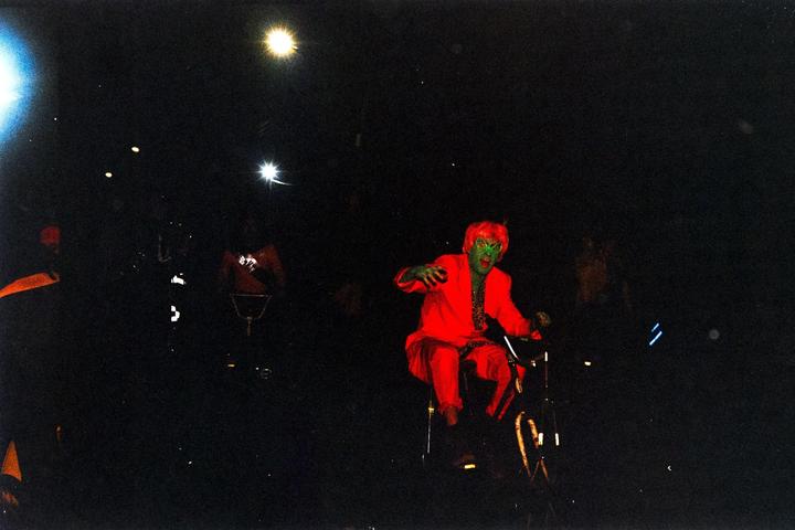 A person with green face paint and red hair, wearing a red suit, rides a bicycle at night.
