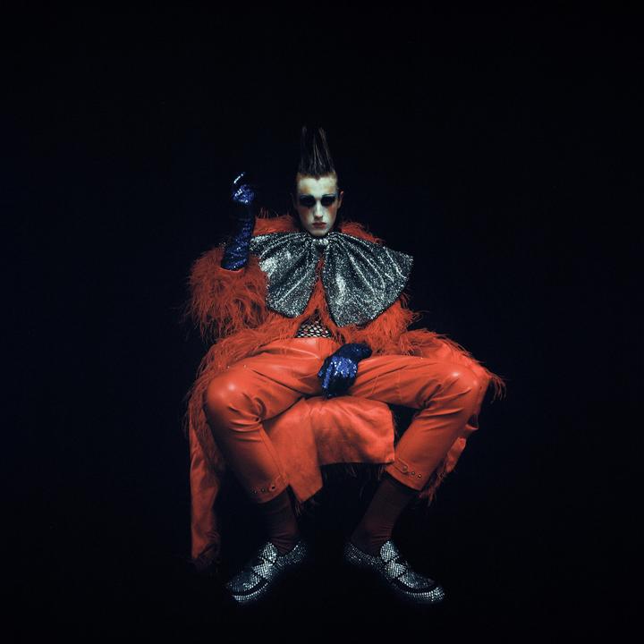 A person in a red feathered coat, red leather pants, and a large sparkly bow sits against a dark background.