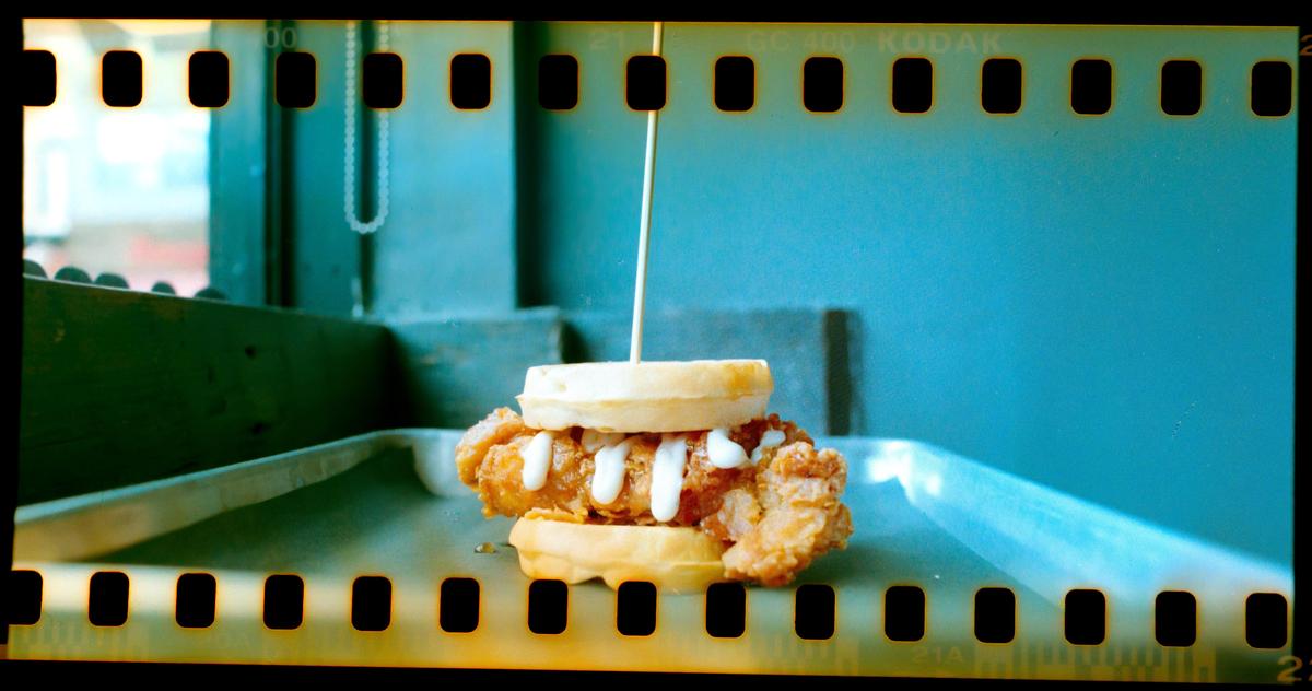A chicken and waffle sandwich with white sauce and a skewer is on a metal tray.