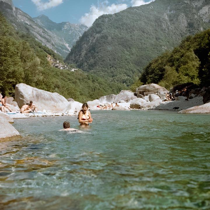 A woman stands in a clear river with a man swimming nearby, surrounded by mountains and people relaxing on the rocky shore.