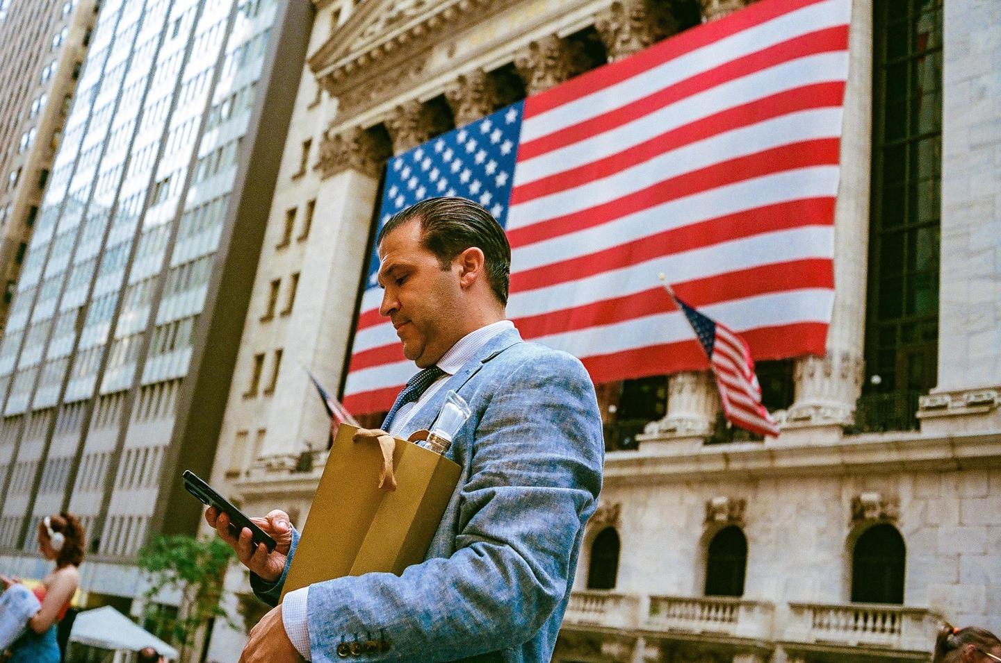 A man in a suit holds a shopping bag and a phone while standing in front of a building with an American flag.