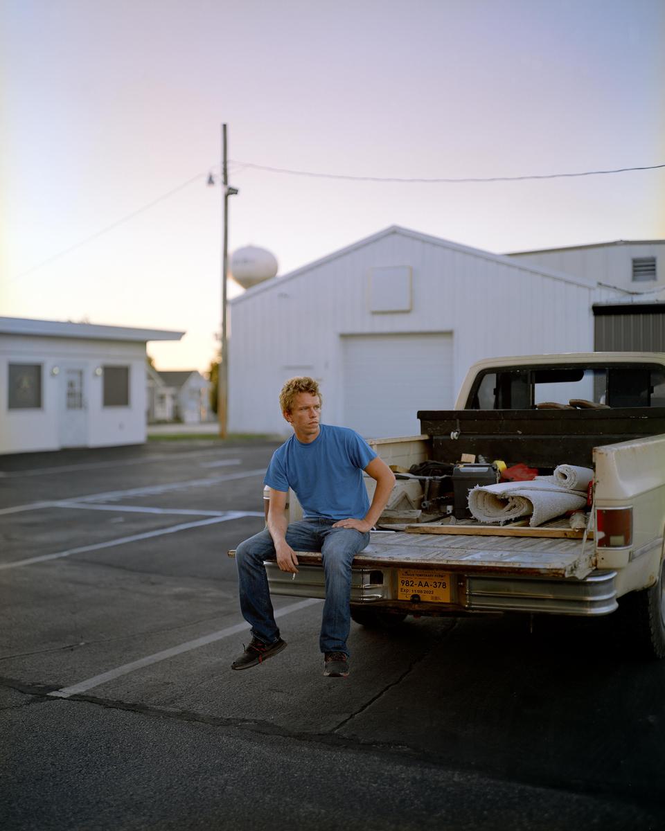 A man sits on the tailgate of a pickup truck in a parking lot with buildings and a water tower in the background.