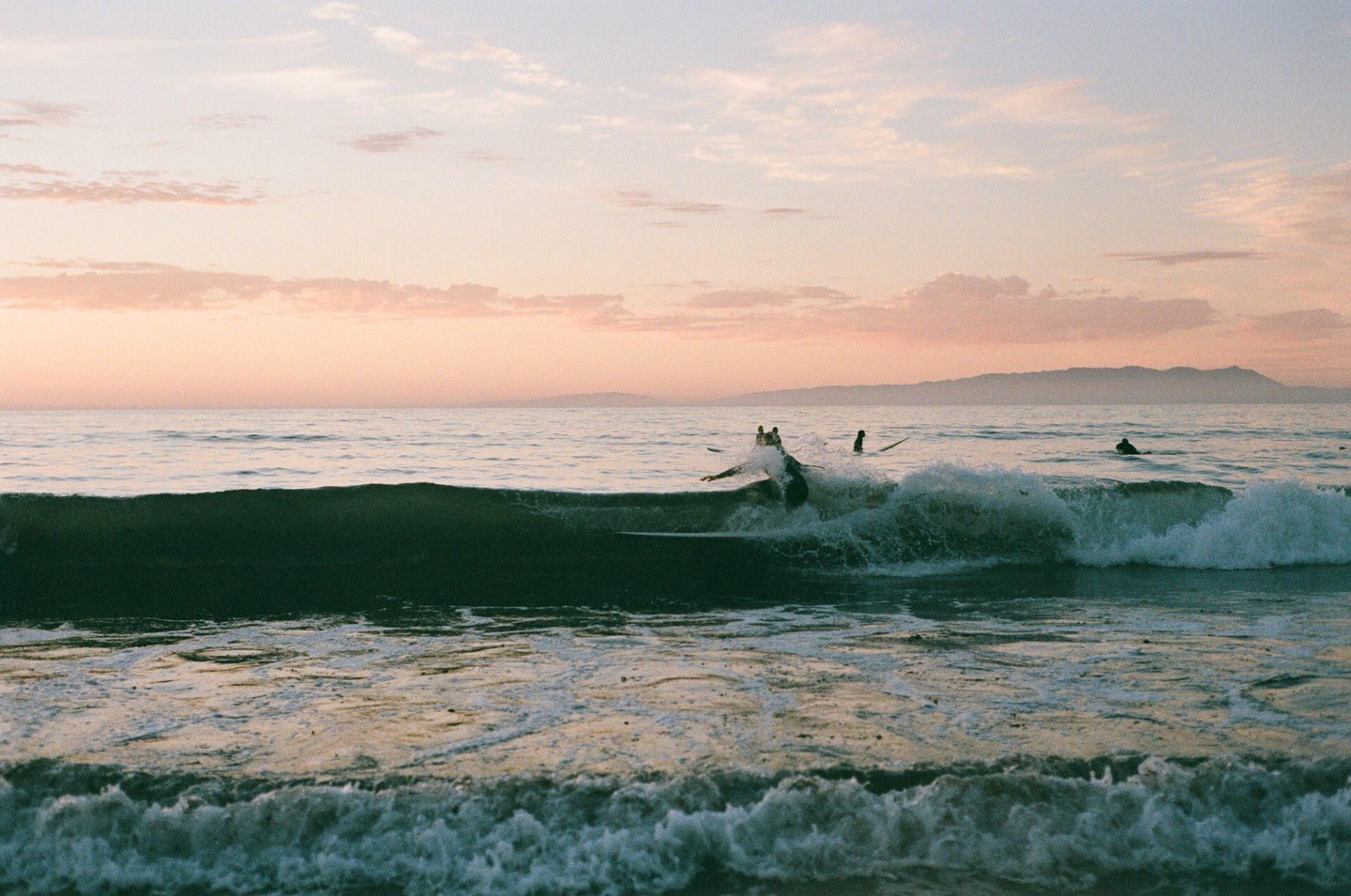 Surfers are in the ocean with waves breaking at sunset.