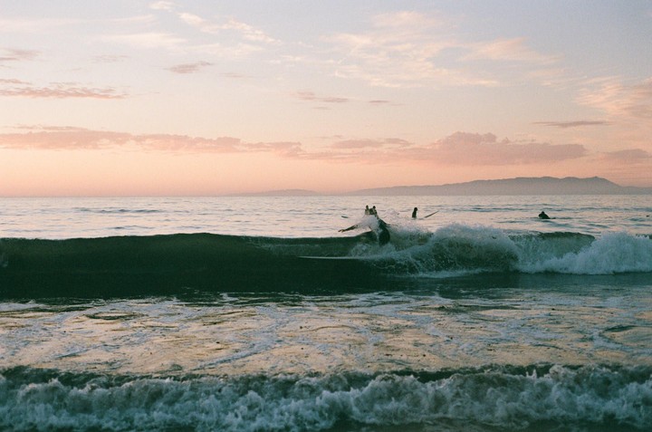 Surfers are in the ocean with waves breaking at sunset.