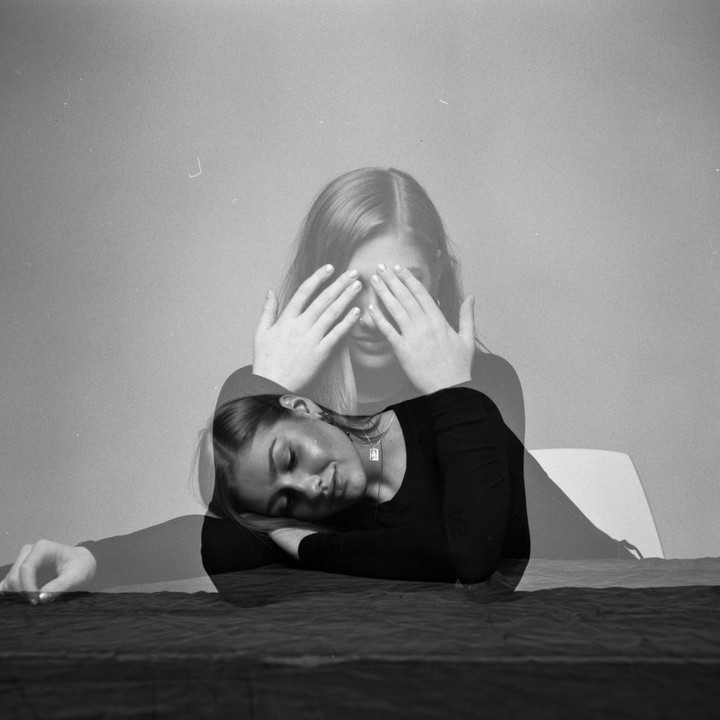 A woman with her eyes covered by her hands is double-exposed over another image of her resting her head on her arm.
