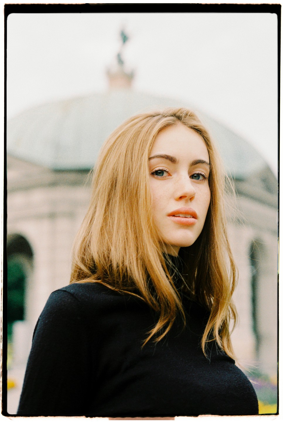 A woman with long hair and a black turtleneck looks at the viewer with a building in the background.