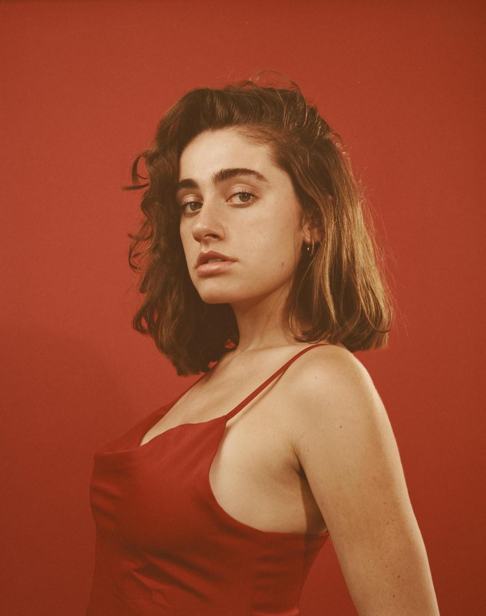 A woman with short, curly hair and a red dress looks directly at the viewer against a red background.