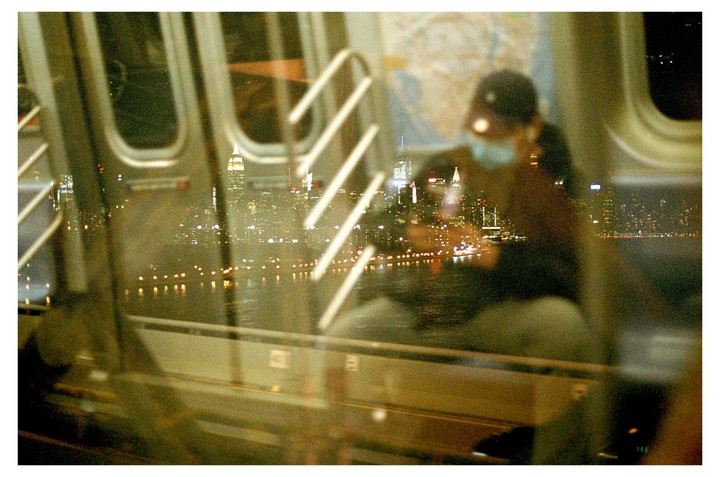 A person wearing a cap and face mask is visible through a window with a reflection of a city skyline at night.