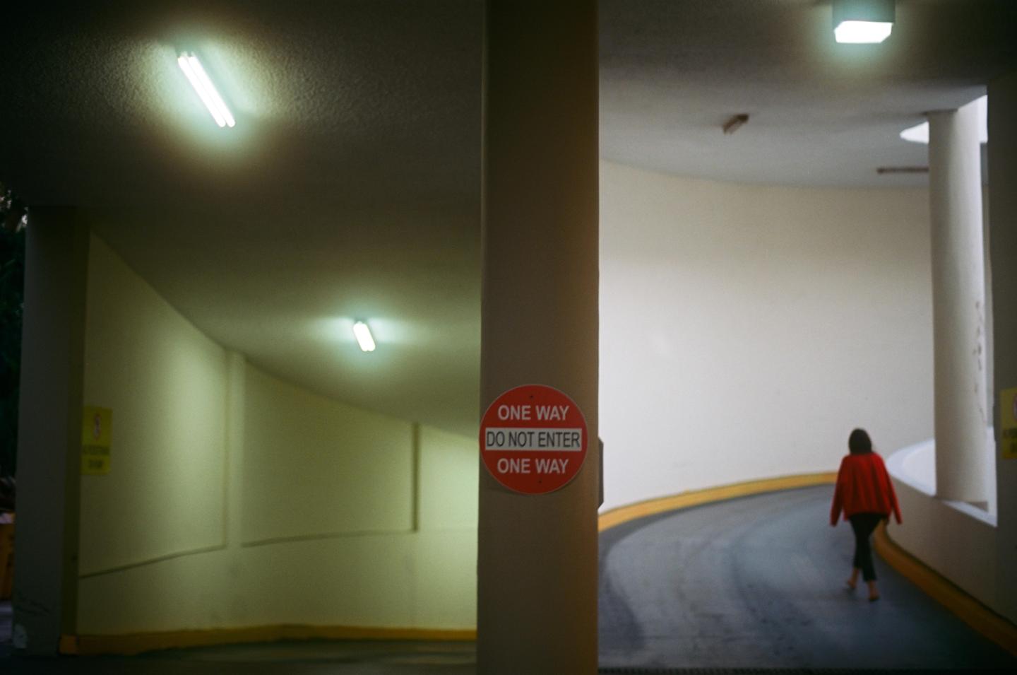 A woman in a red jacket walks away from the camera down a curved ramp in a parking garage.