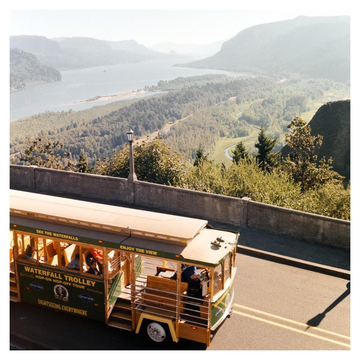 A trolley bus drives on a road overlooking a vast forest and river.