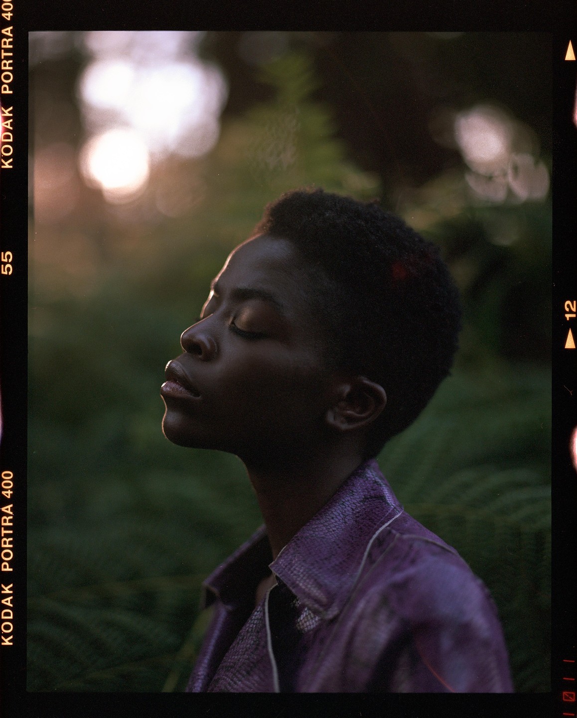 A woman with closed eyes and short hair wears a purple jacket and is backlit by the sun in a forest.