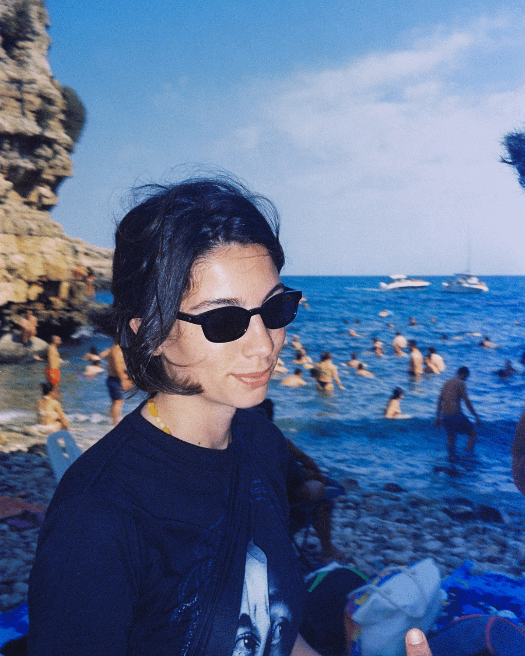 A woman wearing sunglasses and a dark t-shirt sits on a rocky beach with many people swimming in the ocean behind her.