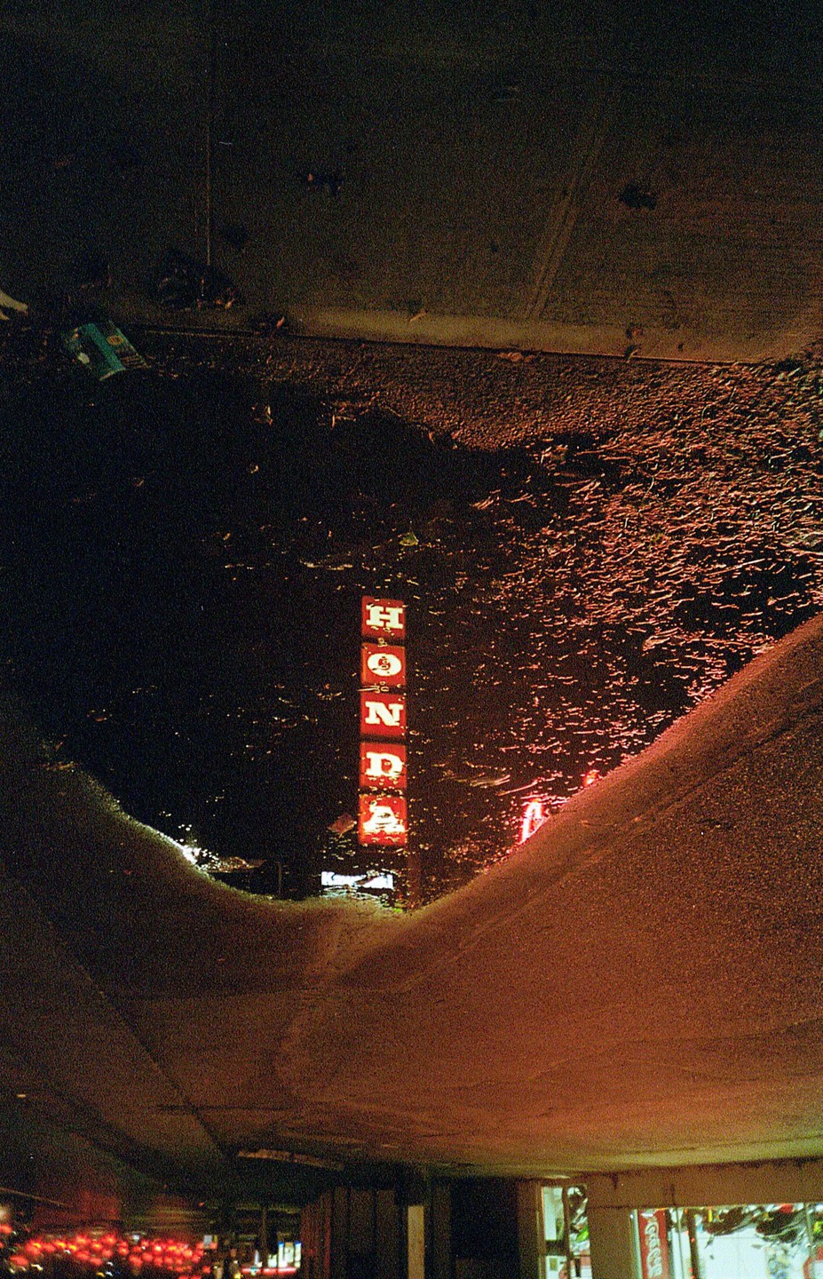 A neon Honda sign is reflected in a puddle on a street at night.