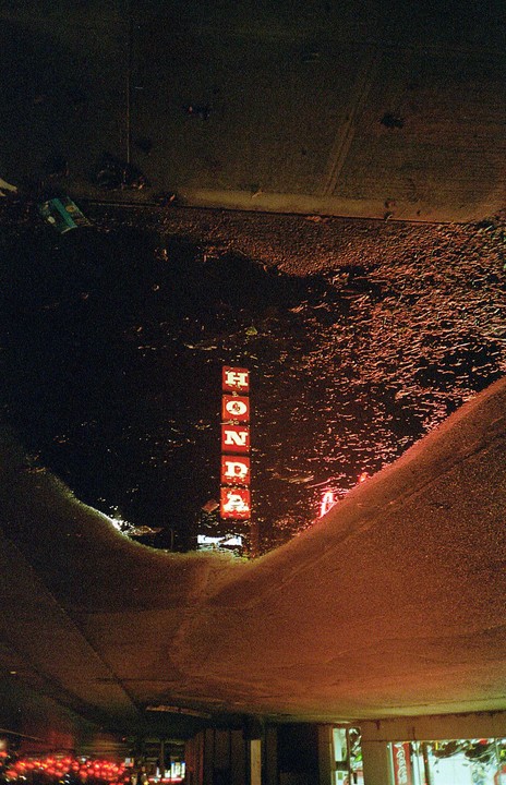 A neon Honda sign is reflected in a puddle on a street at night.