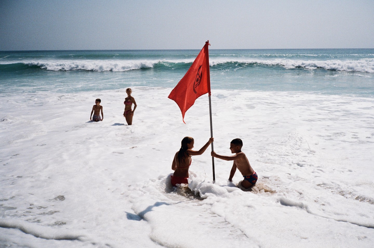 Three people stand in the foamy ocean water, with two children holding a red flag in the foreground and a woman standing further back.
