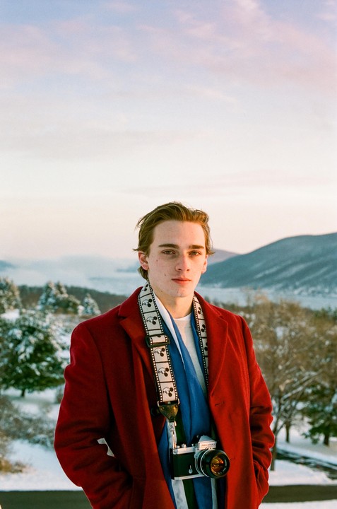 A man in a red coat and blue scarf stands in a snowy landscape with a camera around his neck.