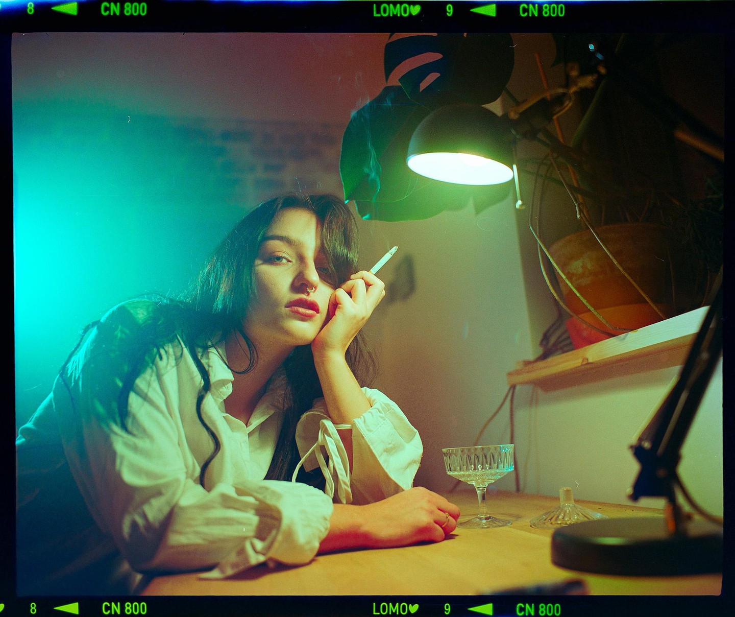 A woman with dark hair and a nose ring holds a cigarette and rests her head on her hand at a table with a glass.