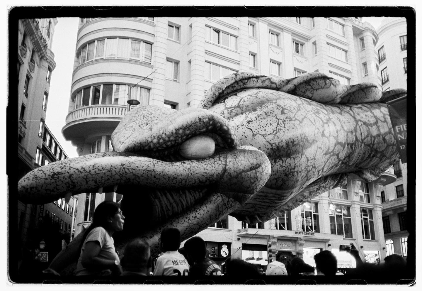 A large dragon float with an open mouth is seen from below, with people watching it on a city street.