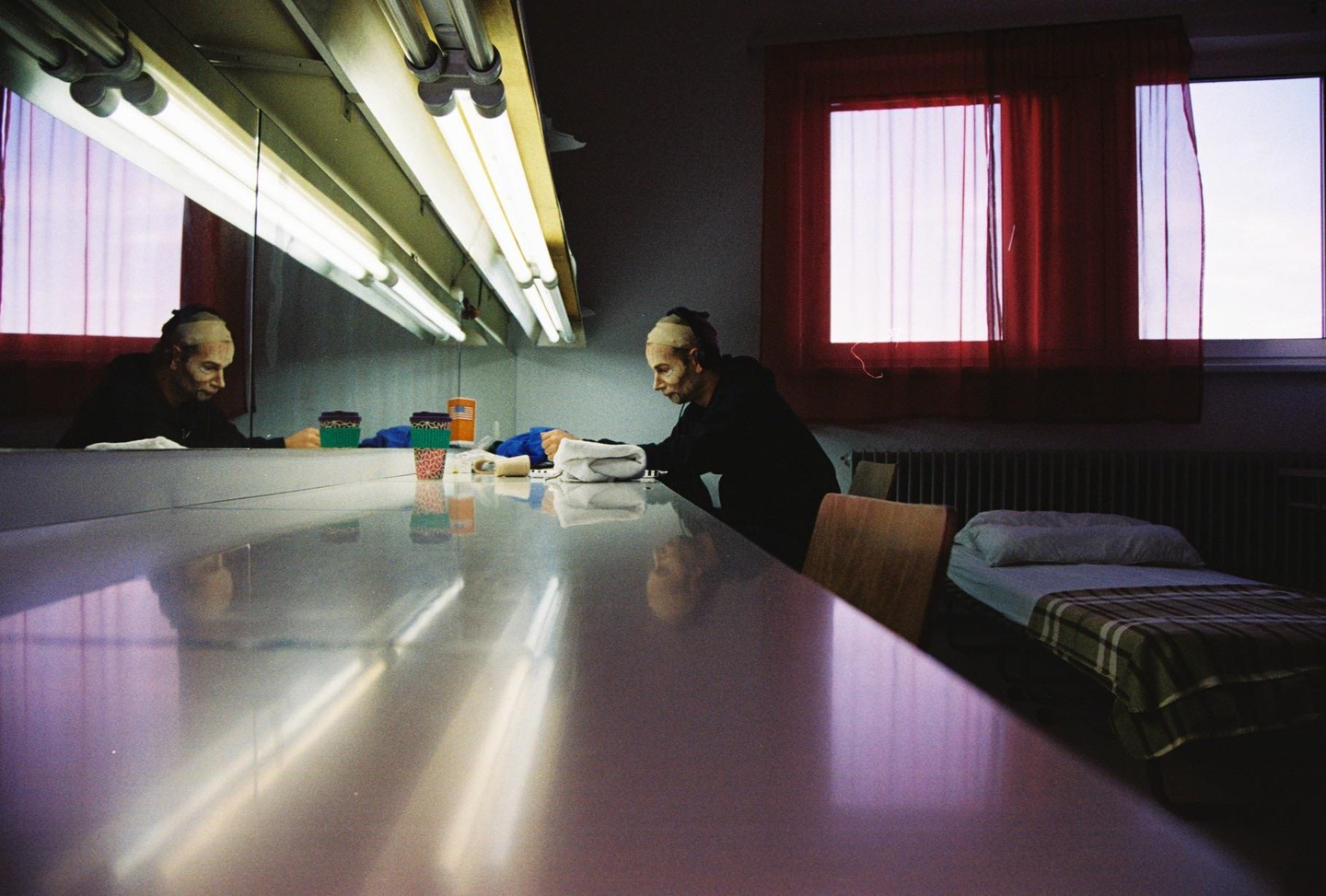 A man with a bald cap sits at a long table with a mirror and fluorescent lights, with a bed and two windows with red curtains in the background.