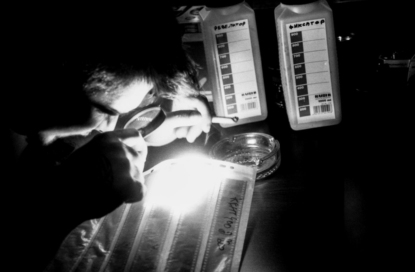 A man is looking at a film strip through a magnifying glass, with two bottles of chemicals and an ashtray on the table.