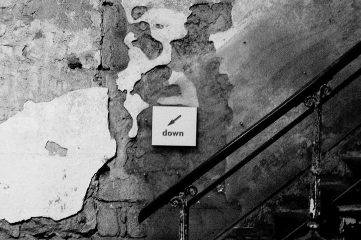 A sign with an arrow pointing down and the word "down" is affixed to a peeling wall next to a metal railing.
