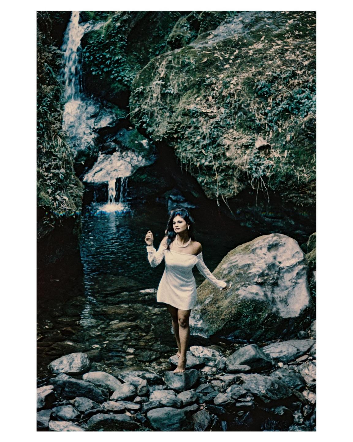 A woman in a white dress stands barefoot on rocks in a stream with a waterfall in the background.
