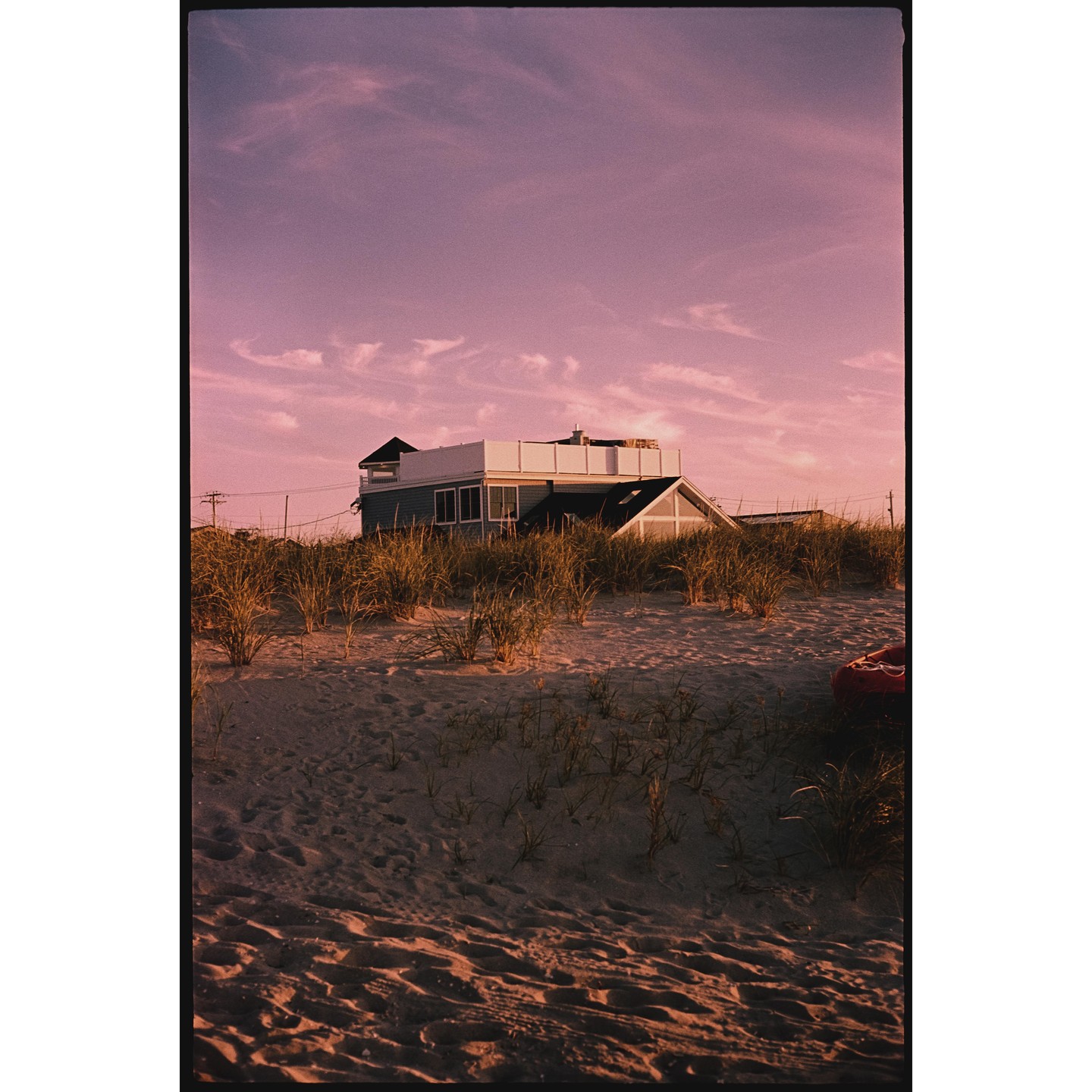 A house with a rooftop deck is visible behind a sandy dune with tall grass under a pink sky.
