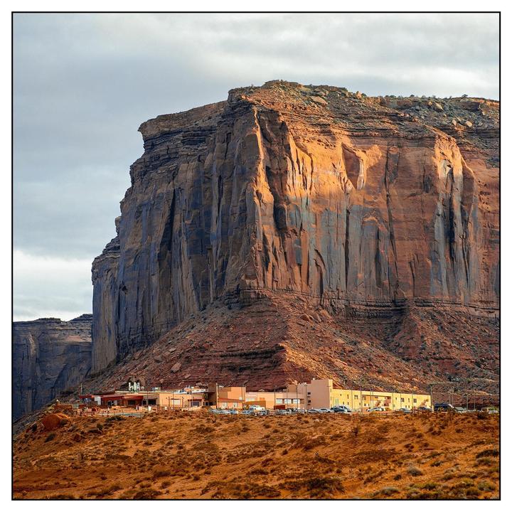 A large rock formation looms over a small settlement of buildings in a desert landscape.