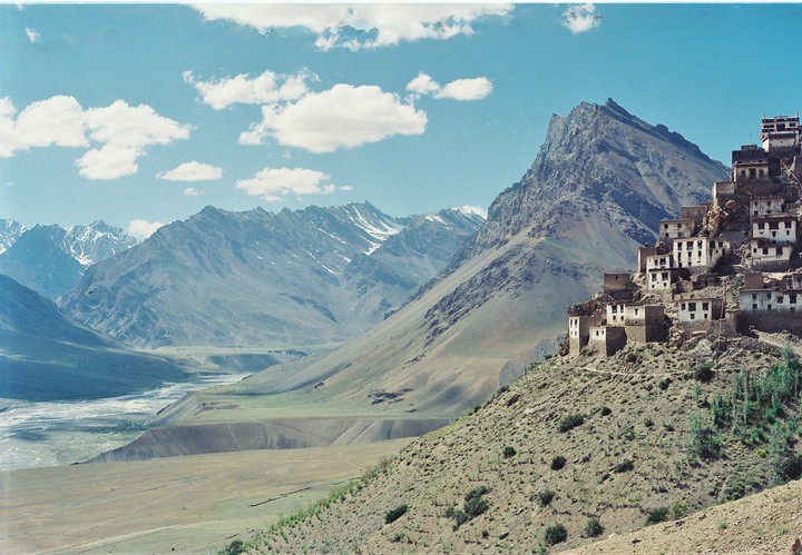 A village built into a steep mountain is visible across a valley with a river and snow-capped mountains under a blue sky with clouds.
