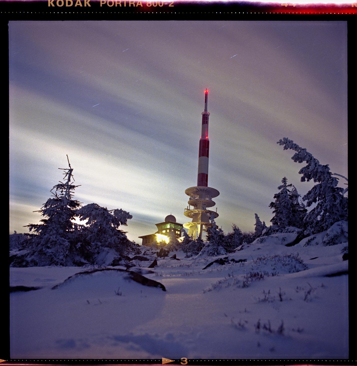 A red and white radio tower stands tall against a cloudy night sky, surrounded by snow-covered trees and ground.