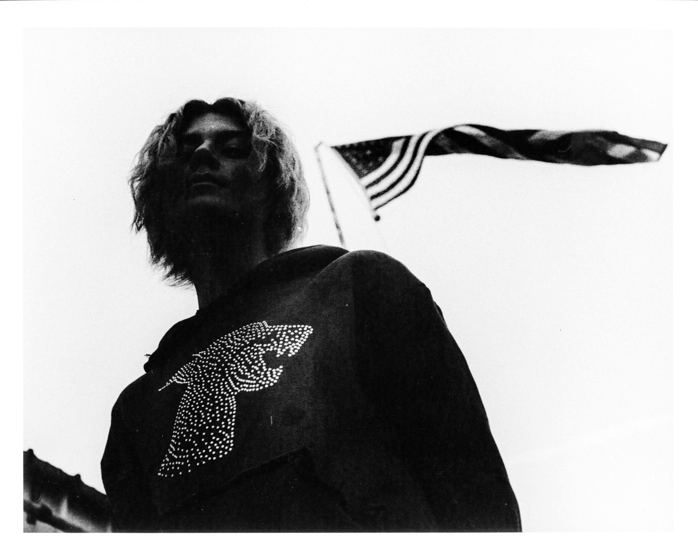 A person with long hair wearing a sweatshirt with a studded animal design looks up at the camera, with an American flag waving in the background.