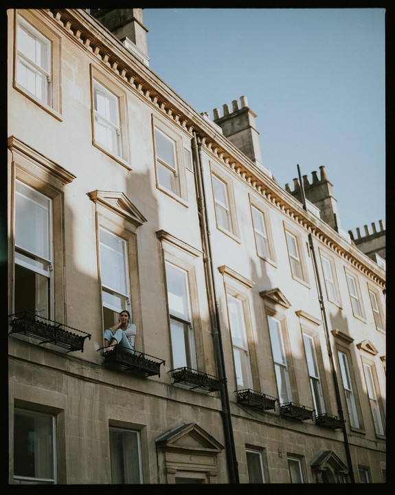 A woman sits on a window ledge of a building with many windows and ornate details under a clear sky.