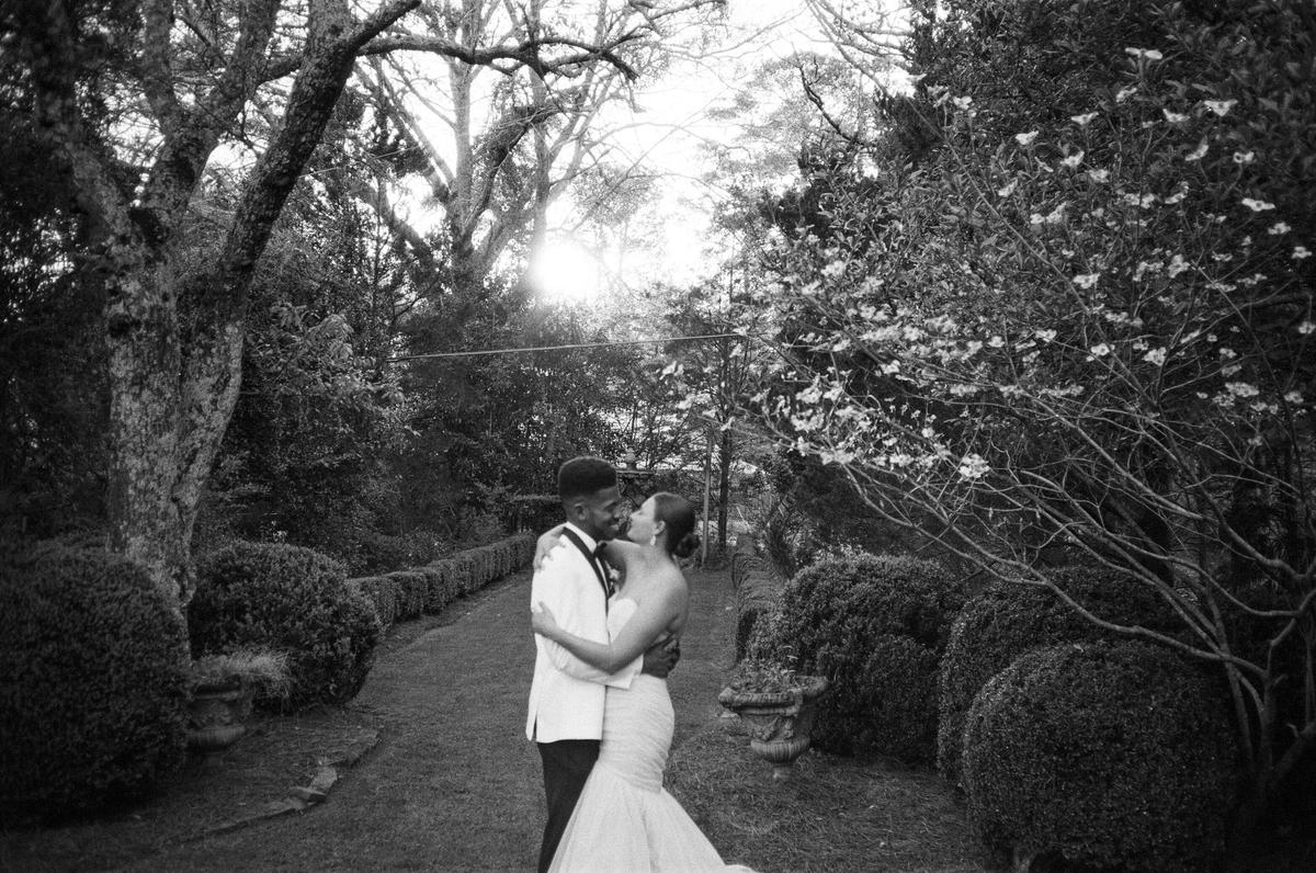 A couple embraces and kisses in a garden with trees and bushes.