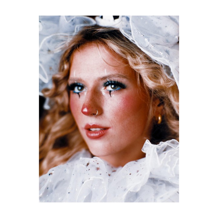A woman with clown makeup and a ruffled collar looks to the side.