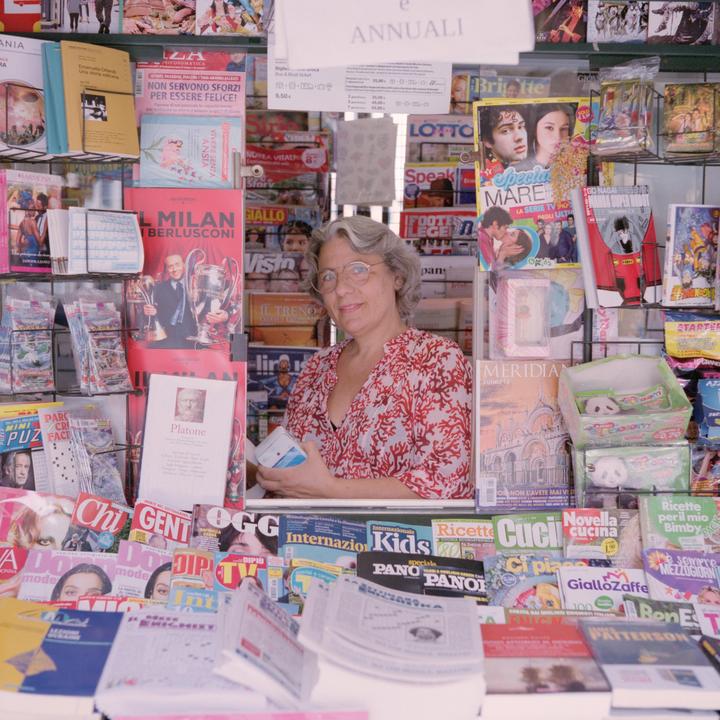 A woman with glasses and a patterned shirt stands behind a counter filled with magazines and newspapers.