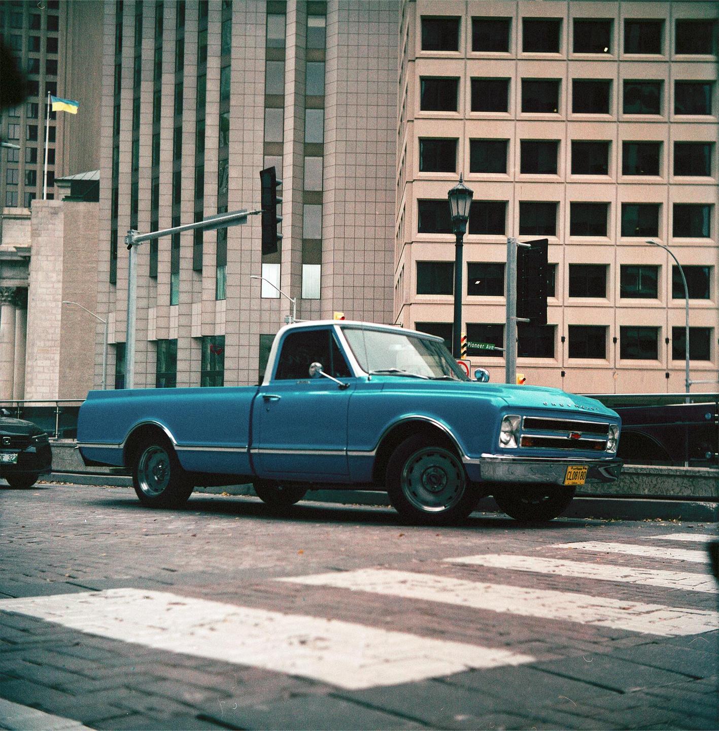 A blue pickup truck is parked on a city street with tall buildings in the background.