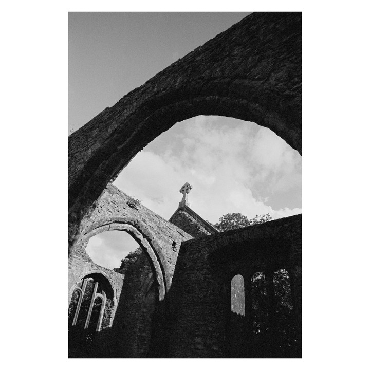 A low-angle, black and white shot of a stone Celtic cross on top of a ruined building, framed by an archway.