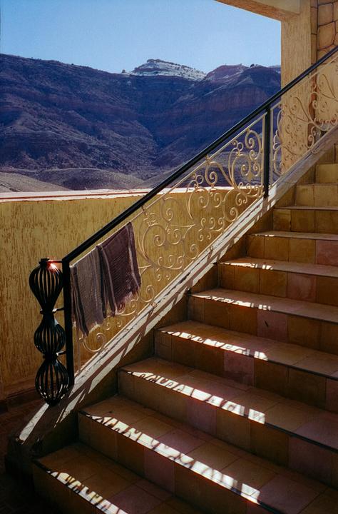 Sunlight casts shadows on a tiled staircase with an ornate railing, overlooking a mountainous landscape.