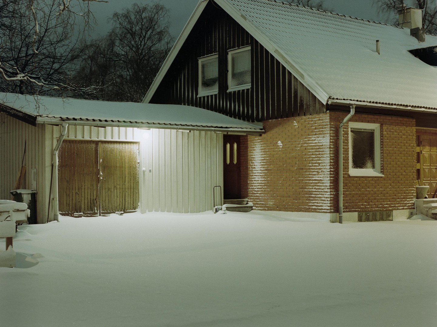 A snow-covered house with a garage is illuminated by a single light at night.