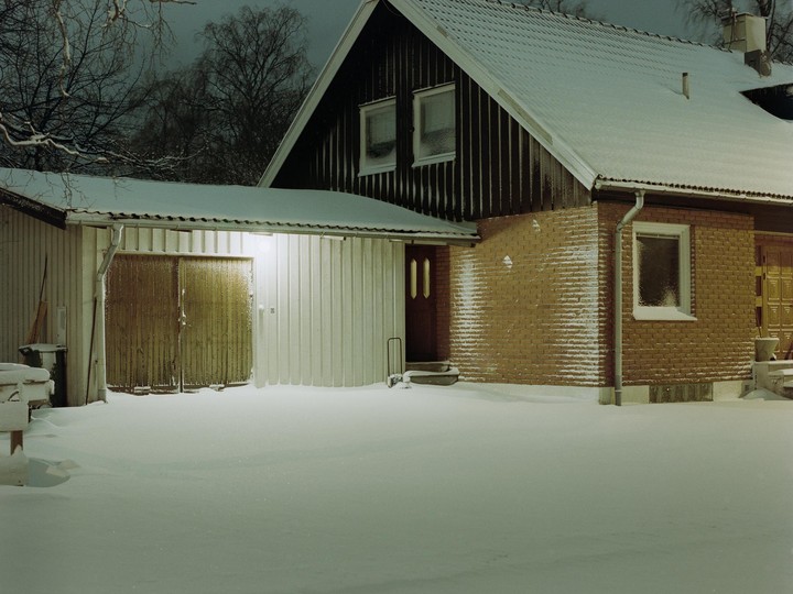 A snow-covered house with a garage is illuminated by a single light at night.