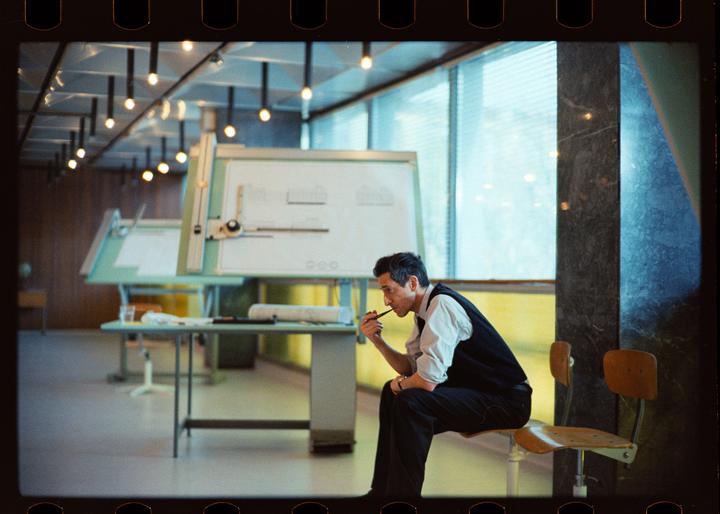 A man sits on a stool in an office with drafting tables and large windows, holding a pipe to his mouth.