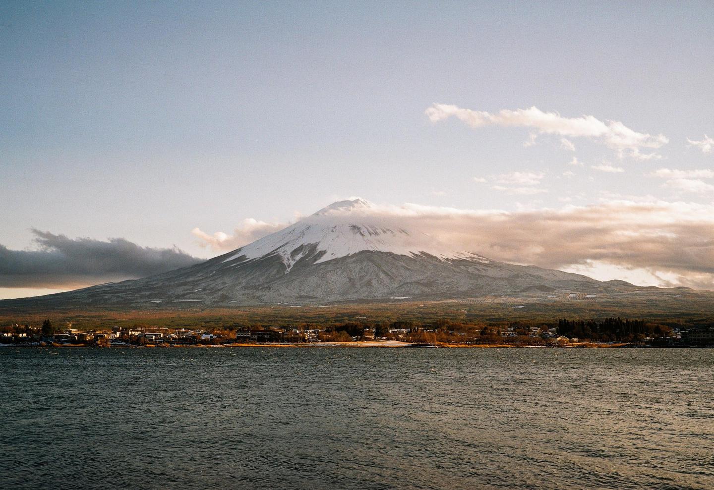 Mount Fuji rises above a town and a body of water under a cloudy sky.