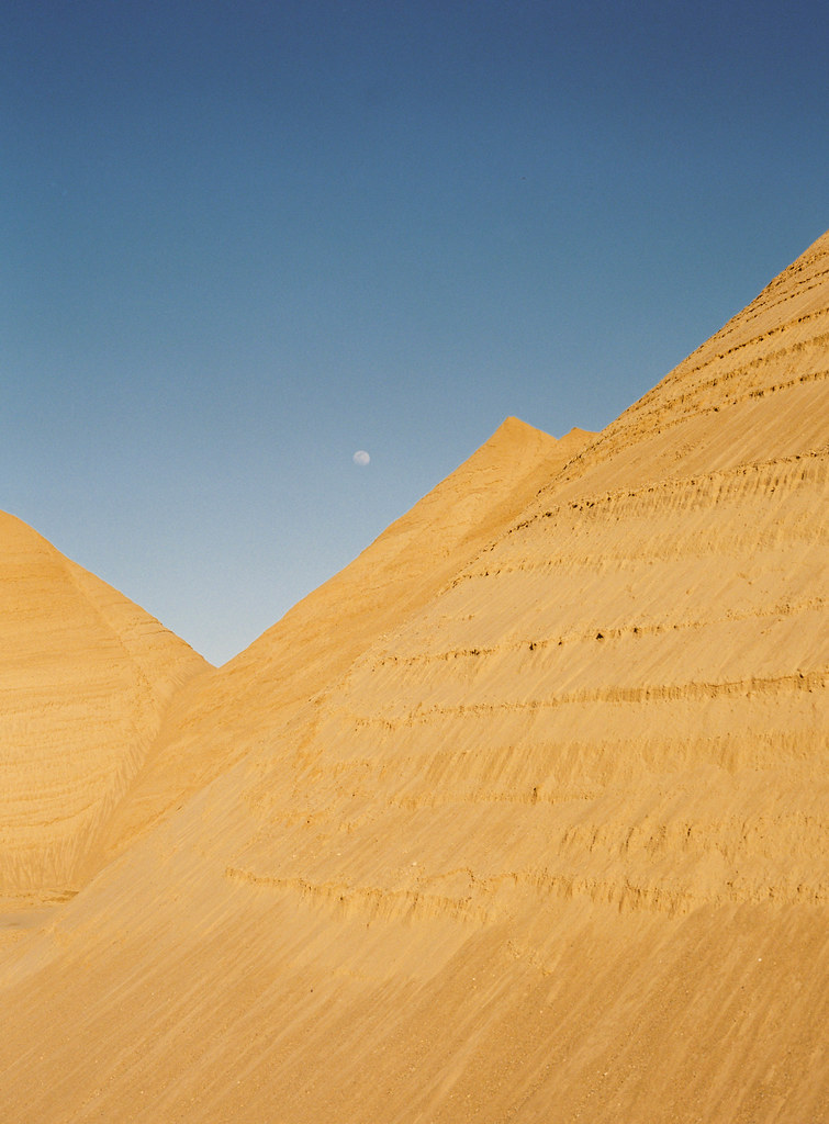A moon is visible in the blue sky above sand dunes.