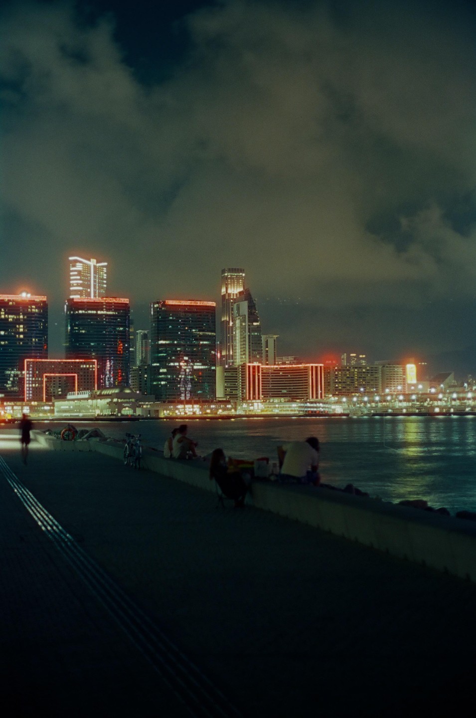 People sit on a wall by the water at night with a city skyline in the background.