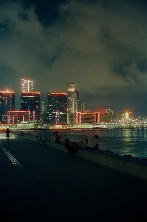 People sit on a wall by the water at night with a city skyline in the background.