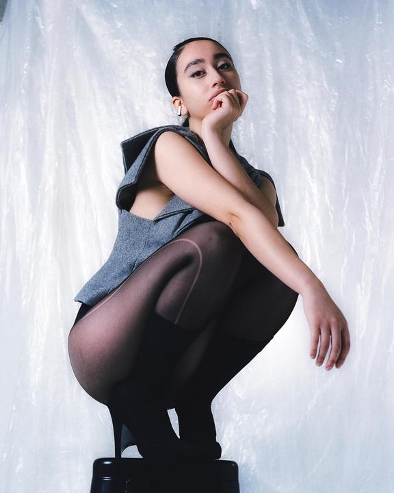 A woman in a gray top and black tights crouches on a black stool against a textured white background.