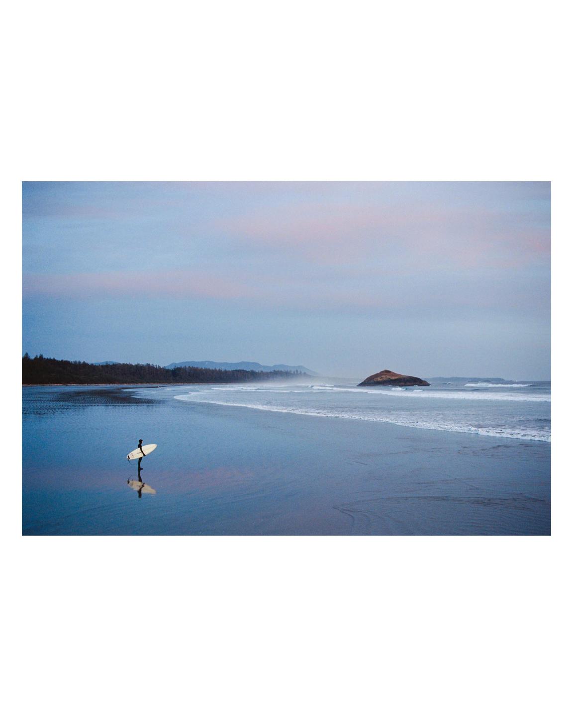 A surfer stands on a beach with a surfboard, reflected in the wet sand, with an island and forest in the distance under a pink and blue sky.
