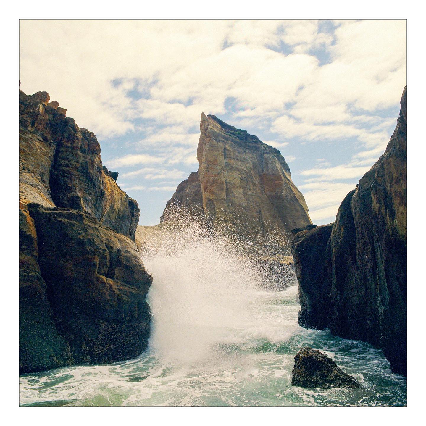 A wave crashes against a rocky cliff face, with a large rock formation in the background under a cloudy sky.