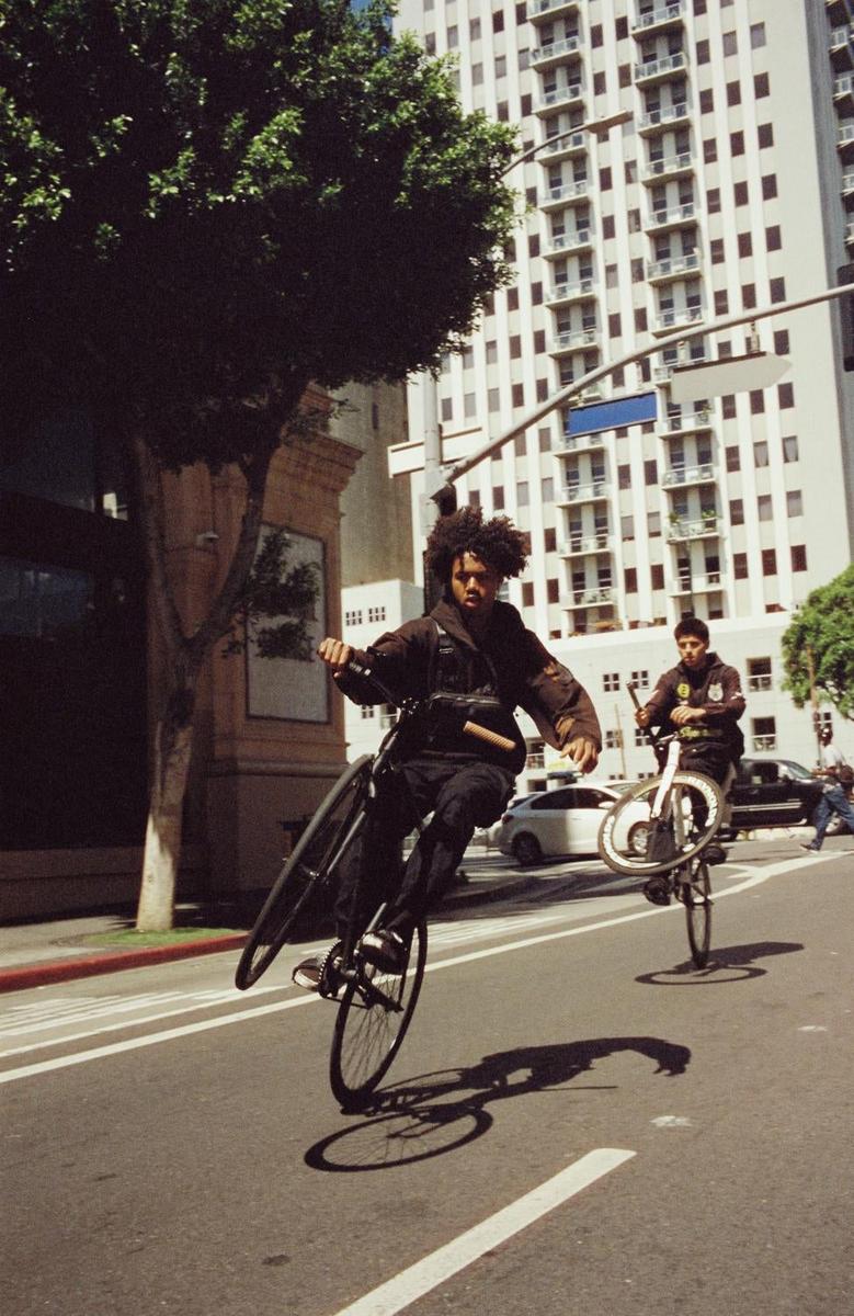 Two men ride bicycles on their back wheels down a city street with a tall building in the background.